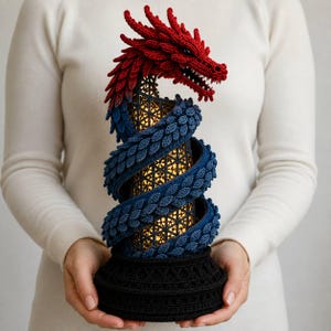 May include: A decorative lamp featuring a crocheted red and blue dragon coiled around a cylindrical, illuminated base with a geometric lattice pattern. The dragon has detailed scales and is mounted on a solid black, textured circular base.