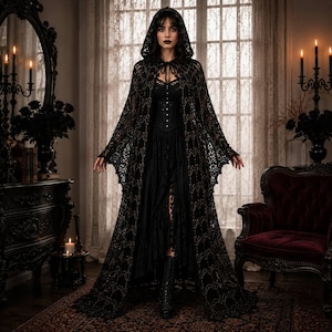 May include: A floor-length black hooded cloak featuring an intricate openwork lace pattern with subtle metallic gold accents. The garment includes a tie closure at the neck and is worn over a black corset-style bodice and a layered skirt with lace trim.
