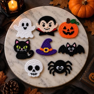 May include: A collection of eight crocheted Halloween-themed appliques including a white ghost, a vampire, an orange pumpkin, a black cat, a purple witch hat, a black bat, a white skull, and a black spider, all arranged on a round marble surface.