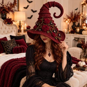 Pode incluir: Um chapéu de bruxa de crochê vermelho-escuro com uma ponta alta e curvada, decorado com rosas de tecido vermelho-escuro, folhas verdes e pequenos ornamentos de olhos embutidos. O chapéu tem uma aba larga e uma textura de fio tricotado.