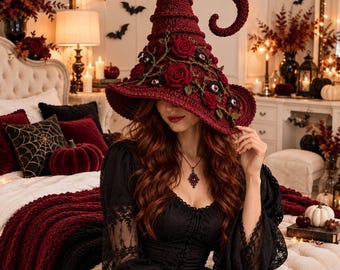 Puede incluir: Un sombrero de bruja de ganchillo en color rojo oscuro con una punta alta y curvada, decorado con rosas de tela rojo oscuro, hojas verdes y pequeños adornos de ojos incrustados. El sombrero tiene un ala ancha y una textura de hilo.