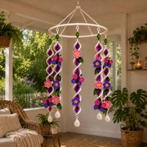 May include: A hanging mobile featuring a circular white frame with five dangling crochet strands. Each strand is a spiral of white, purple, and pink yarn, decorated with crocheted butterflies, flowers, and green leaves, ending in a small white bead.