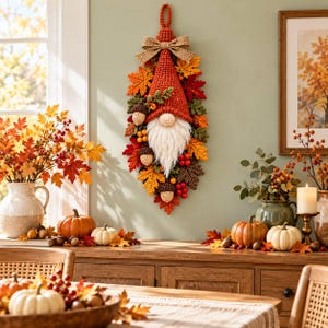 Könnte beinhalten: Ein vertikaler Wandbehang mit einem gehäkelten Wichtel, der eine hohe rote Mütze, einen weißen Bart und eine Schleife aus Jute trägt. Der Wichtel ist von einer herbstlichen Dekoration aus gehäkelten Blättern, roten Beeren und braunen Eicheln umgeben.