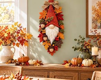 Könnte beinhalten: Ein vertikaler Wandbehang mit einem gehäkelten Wichtel, der eine hohe rote Mütze, einen weißen Bart und eine Schleife aus Jute trägt. Der Wichtel ist von einer herbstlichen Dekoration aus gehäkelten Blättern, roten Beeren und braunen Eicheln umgeben.
