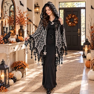 May include: A black hooded crochet cape featuring a white spiderweb pattern, adorned with dangling black beads and silver-toned star and crescent moon charms. The garment has a tie closure at the neck with matching moon-shaped accents.