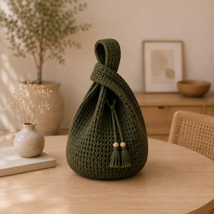 Può includere: Una borsa all'uncinetto color verde oliva scuro con un design a manico annodato strutturato. La borsa presenta un motivo intrecciato testurizzato e include due cordini pendenti rifiniti con piccole perline di legno e nappe verdi abbinate.