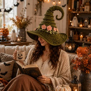Puede incluir: Un sombrero de bruja de ganchillo en color verde oliva oscuro con una punta alta y curvada, y un ala ancha adornada con una banda de pequeñas flores de ganchillo rosa claro y hojas verdes. El sombrero presenta un patrón de punto acanalado.