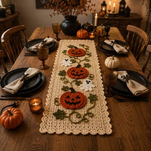 Puede incluir: Un camino de mesa rectangular de ganchillo color crema con tres calabazas naranjas y dos fantasmas blancos, decorado con enredaderas y hojas verdes. El camino tiene un borde festoneado y está sobre una mesa de comedor de madera.