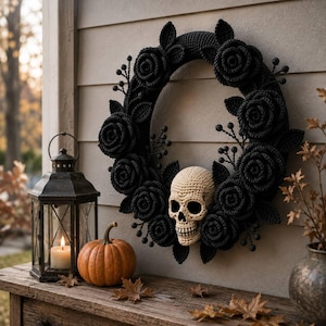 May include: A circular wreath crafted from black crocheted roses and leaves, featuring a central cream-coloured crocheted skull. The decorative piece is adorned with small black berry-like accents and is displayed against a horizontal panelled wall.