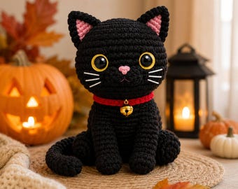 Könnte beinhalten: Ein schwarzes, gehäkeltes Katzen-Stofftier mit großen gelben Augen, einer kleinen rosa Nase, weißen gestickten Schnurrhaaren und rosa Innenohren. Die Katze trägt ein rotes Halsband mit einer kleinen goldenen Glocke in der Mitte und sitzt.