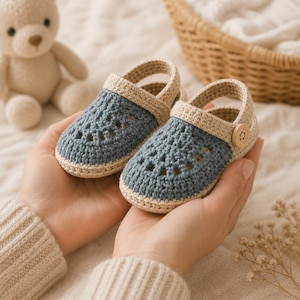 May include: A pair of baby shoes crafted from blue and beige yarn. The shoes feature a crochet pattern with openwork detailing on the toe area, a beige strap across the top, and a small wooden button on the side of each shoe for a secure fit.