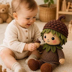 Puede incluir: Una muñeca de ganchillo hecha a mano con forma de personaje de bellota, con un gorro texturizado de color marrón oscuro, un cuello de hojas verdes y una cara de color tostado claro con mejillas sonrosadas. La muñeca sostiene una bellota.