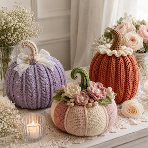 May include: Three crocheted pumpkins in lavender, orange, and a mix of pink and cream. The lavender pumpkin features a white lace bow, the orange one has a brown stem and white ruffled collar, and the pink and cream one is adorned with crocheted flowers.