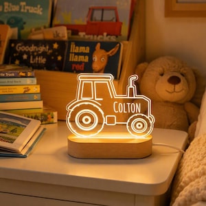 Puede incluir: Una luz nocturna LED brillante con forma de tractor, que presenta el nombre Colton grabado en el centro. La luz está montada sobre una base de madera clara y lisa, y se alimenta mediante un cable blanco que sale del lateral de la base.