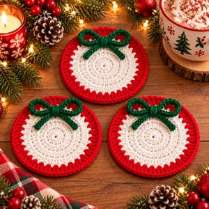 May include: Three circular crochet coasters featuring a white centre, a red outer border, and a small green yarn bow attached to the top edge. The coasters are arranged on a wooden surface, each displaying a consistent handmade textile design.
