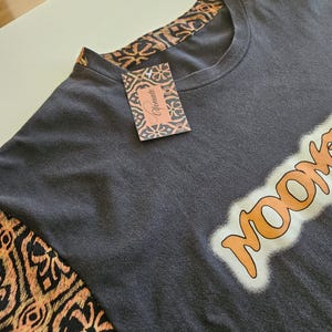 Puede incluir: Una camiseta gris carbón con detalles de tela estampada en naranja y negro en el cuello y las mangas. La prenda muestra la palabra "Noom" en letras naranjas con un borde blanco e incluye una etiqueta de marca a juego con el texto "Yomate".