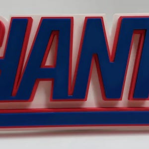 May include: A three-dimensional wall sign featuring the word GIANTS in bold blue letters with a red outline, mounted on a white rectangular base. A horizontal blue bar with a red outline is positioned directly underneath the text.