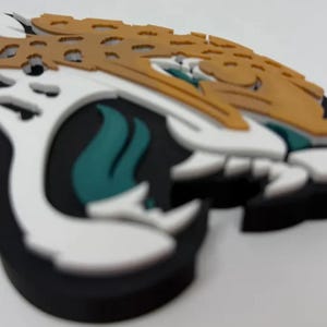 May include: A three-dimensional layered wall art piece depicting a stylized jaguar head logo. The design features a black base, white accents, a teal tongue, and a gold-colored top layer with cut-out details, creating a multi-layered relief effect.