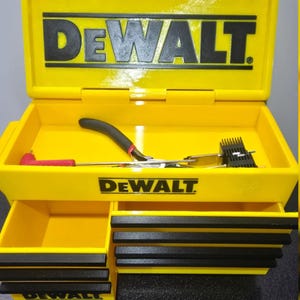 May include: A bright yellow DeWalt tool chest with a hinged lid and multiple drawers. The DeWalt logo is printed in black on the lid, the front panel, and the bottom drawer. Inside the open top compartment are a pair of pliers and an L-shaped tool.