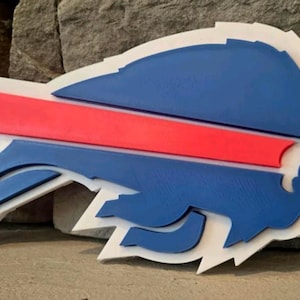 May include: A layered wall sign featuring the Buffalo Bills logo. The design consists of a blue bison silhouette, a bright red horizontal streak, and a white backing layer, creating a three-dimensional effect with distinct stacked components.