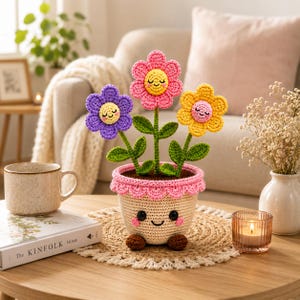 Pode incluir: Uma planta em vaso de crochê com três flores sorridentes em roxo, rosa e amarelo com caules e folhas verdes. O vaso bege tem uma borda rosa festonada, olhos pretos bordados, um pequeno sorriso e dois pequenos pés castanhos na base.