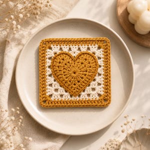 May include: A square crochet coaster featuring a mustard yellow heart motif centered within a white and mustard yellow geometric border. The textile piece is crafted with a textured yarn pattern, showcasing a distinct heart shape surrounded by a square frame.