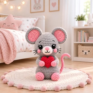 Pode incluir: Um peluche de rato feito em croché cinzento com grandes orelhas redondas com centros cor-de-rosa, um pequeno nariz cor-de-rosa, olhos pretos bordados e bochechas cor-de-rosa. O rato segura um coração de croché vermelho vivo e tem uma cauda rosa curva.