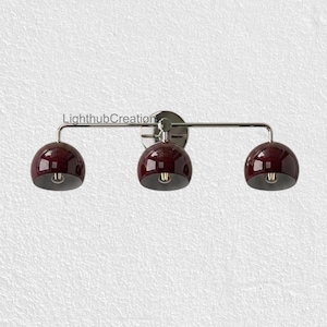 Peut inclure: Une applique murale à trois lumières dotée d'une barre horizontale en chrome poli et de trois abat-jours en forme de dôme bordeaux foncé brillant. Le luminaire porte le texte LighthubCreations au-dessus de la lumière centrale.