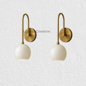 Può includere: Una coppia di applique da parete con piastra circolare in ottone, braccio curvo in ottone e paralume sferico in vetro bianco. Il testo LightHubCreations è visibile al centro. Questi apparecchi offrono un design pulito e simmetrico.