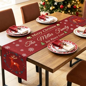 May include: A deep red table runner featuring white snowflake patterns, a reindeer wreath design, and the printed text Merry Christmas, The Miller Family, and Joy-Peace-Love. The runner is displayed on a wooden table with matching napkins.