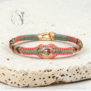 May include: A bracelet featuring a central rectangular clear stone surrounded by small multicolored gemstones, set in a gold-toned frame. The band is constructed from braided olive green and bright orange cord, finished with a gold-toned metal clasp.