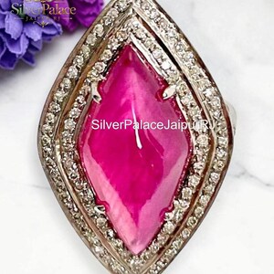 May include: A marquise-shaped ring featuring a large, smooth, vibrant pink center stone surrounded by a double halo of small, clear, sparkling stones set in a metallic frame, with the text SilverPalaceJaipurRJ visible.