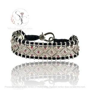 May include: A bracelet featuring a black cord base with a silver-toned metal frame. The frame is composed of diamond-shaped segments encrusted with small clear stones and accented with small round red stones. It includes a silver-toned hook clasp.