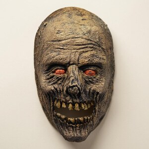 May include: A full-face mask featuring a textured, weathered brown surface with deep wrinkles and sunken features. The mask has an open mouth revealing yellowed teeth and prominent, glowing orange eyes set deep within the eye sockets.