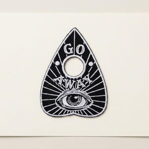 May include: A black and white embroidered patch shaped like a planchette. The design features a central circular cutout, the text GO AWAY in white embroidery, and a detailed eye graphic at the bottom, all surrounded by radiating white lines.