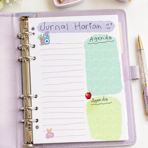 Può includere: Un raccoglitore a sei anelli color lavanda contenente una pagina di agenda bianca con il testo Jurnal Harian, due sezioni etichettate come Agenda, una piccola illustrazione di una fotocamera, un'icona di mela rossa e carta a righe viola.