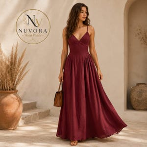 May include: A floor-length, deep burgundy maxi dress with a V-neckline and thin spaghetti straps. The dress features a fitted bodice and a flowing, pleated skirt. A small brown handbag is held at the side. The logo NUVORA WearFinds is visible.