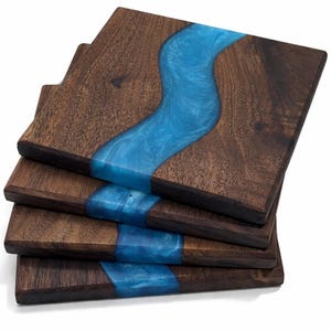 May include: A stack of four square coasters made from dark brown wood with a prominent, wavy, translucent blue resin inlay running diagonally across the centre of each piece, creating a river-like pattern on the surface of the wood.