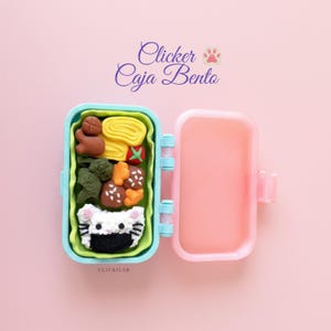 Puede incluir: Una caja bento de color azul claro y rosa que contiene alimentos en miniatura, incluyendo una bola de arroz blanca con forma de gato, brócoli, huevo y trozos de carne. El texto "Clicker Caja Bento" y un icono de huella son visibles arriba.