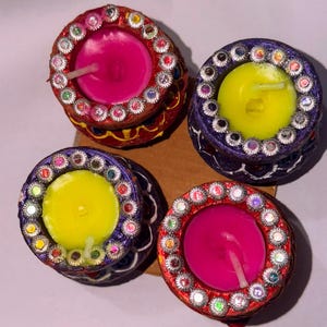 May include: Four round clay diya lamps arranged in a square. Two are red with pink wax, and two are purple with yellow wax. Each lamp features a rim decorated with small, reflective, multi-colored circular embellishments and a single white wick.