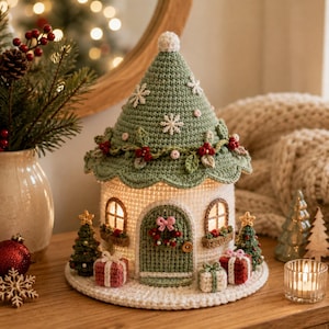 Könnte beinhalten: Ein gehäkeltes Weihnachtshaus als Dekoration mit einem salbeigrünen, kegelförmigen Dach, weißen Schneeflocken-Applikationen und einer roten Beerengirlande. Die weißen Wände haben zwei beleuchtete Fenster, eine grüne Bogentür und kleine Geschenke.