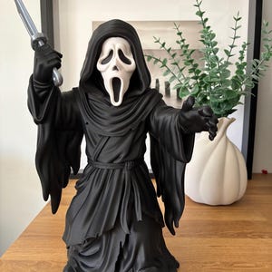Peut inclure: Une figurine Ghostface avec un masque blanc aux ouvertures noires pour les yeux et la bouche, vêtue d'une robe à capuche noire avec une taille nouée. La figurine tient un couteau argenté dans sa main droite et tend la main gauche.