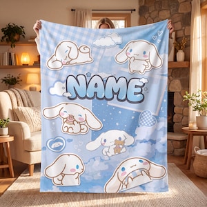May include: A light blue fleece blanket featuring a repeating pattern of the white Cinnamoroll character in various poses, including sleeping, eating, and holding a teddy bear. The word NAME is printed in large, bold, blue bubble letters.