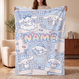 Peut inclure: Une couverture en polaire bleu clair ornée d'un collage de personnages Cinnamoroll, de cadres photo et d'une zone de texte centrale aux couleurs arc-en-ciel portant l'inscription NAME. Le motif inclut des marguerites, des carreaux vichy.
