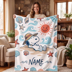 May include: A white fleece blanket featuring a cartoon dog wearing a straw hat while relaxing on a sandy beach. The design includes a smiling orange sun, blue ocean waves, and various colorful seashells and starfish, with the word NAME printed.