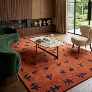 May include: A rectangular area rug featuring a burnt orange base color with a repeating pattern of dark navy blue abstract leaf-like motifs. The rug is positioned on a dark wood floor, partially underneath a coffee table and a curved green sofa.