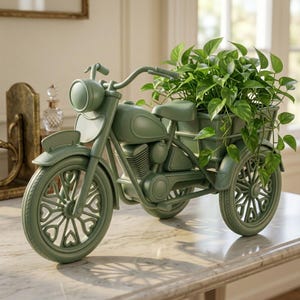 Pode incluir: Um vaso em forma de motocicleta na cor verde sálvia, com farol dianteiro, guidão e uma caçamba traseira preenchida com uma planta pothos de folhagem verde. As rodas apresentam um padrão repetitivo de raios em formato de coração no aro.