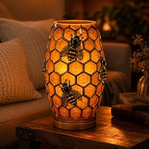 May include: A cylindrical table lamp with a warm amber glow, featuring a raised honeycomb pattern across the surface. Three dark, detailed bee figures are attached to the exterior, contrasting with the translucent, textured yellow glass.