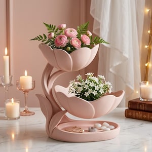 Könnte beinhalten: Ein gestufter, hellrosa, blumenförmiger Keramikständer mit zwei schalenartigen Ebenen und einer Basisschale. Die obere Schale enthält rosa Ranunkeln und Grün, die mittlere Schale kleine weiße Blumen und die Basisschale enthält Schmuck.