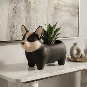 Könnte beinhalten: Ein mattschwarzer und beigefarbener Keramikübertopf in Form eines Corgi-Hundes. Der Übertopf hat einen glatten, abgerundeten Körper mit aufrechten Ohren und einer spitzen Schnauze und enthält eine kleine grüne Bogenhanfpflanze mit panaschierten Blättern.